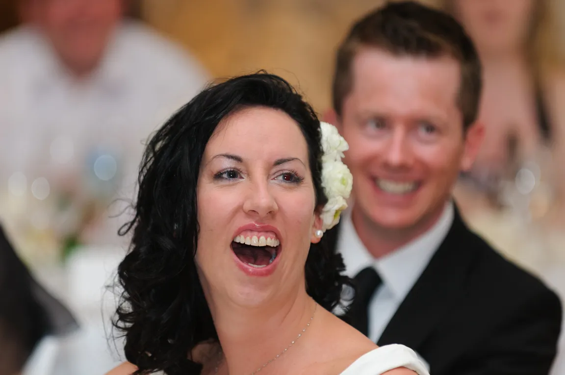 Bride with dark curly hair and white flower smiling and laughing with groom in a black suit smiling behind her.