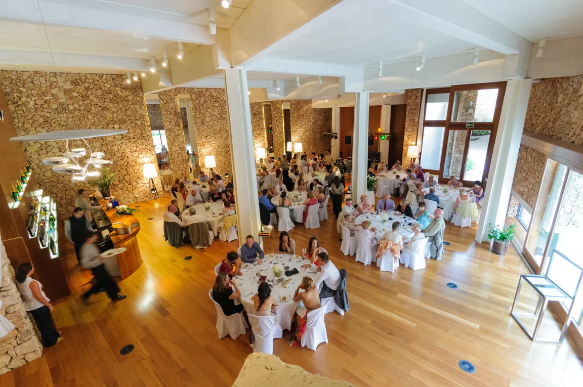 Indoor dining area with wooden floors and stone walls filled with guests seated at round tables covered in white tablecloths.
