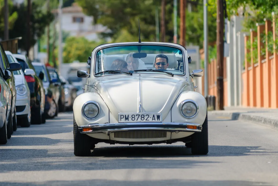 Front view of a white vintage convertible car with three people inside driving on a street lined with parked cars and trees.