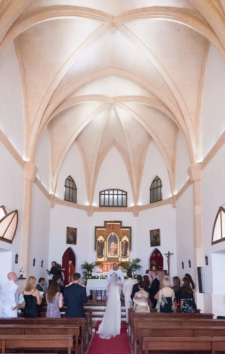 Wedding ceremony inside a chapel with vaulted ceiling, bride and groom standing at the altar with priest and guests seated on wooden benches.