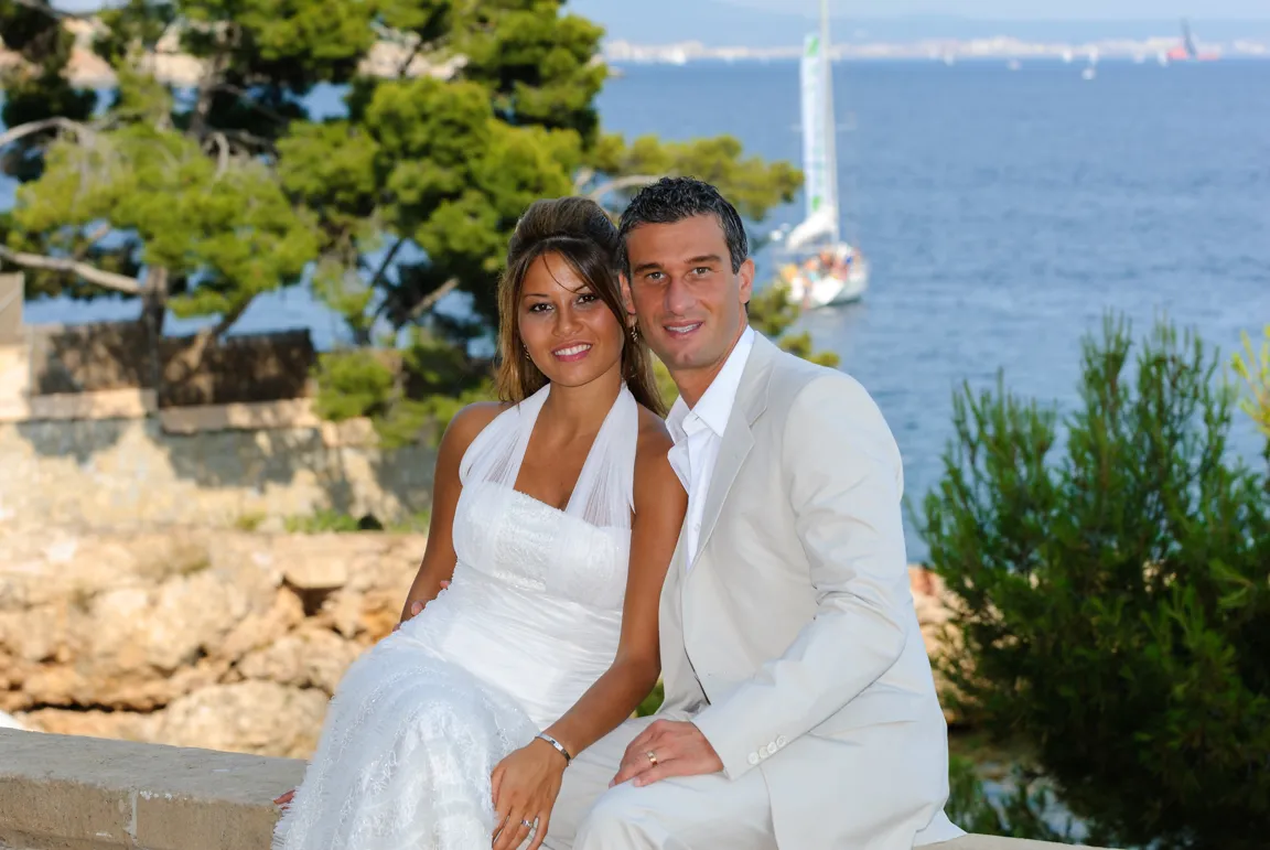 Bride in white dress and groom in light suit sitting together outdoors near water with sailboats in the background.