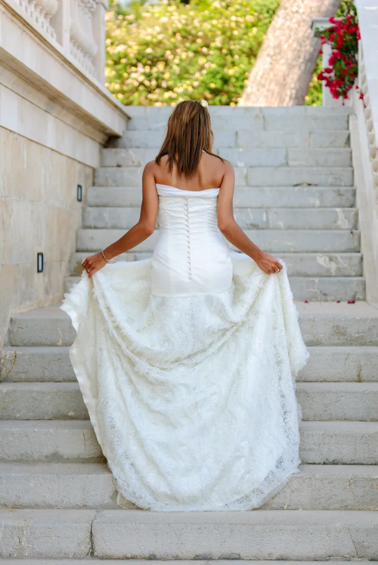 Woman in a strapless white wedding dress holding the train while walking up stone steps outdoors.