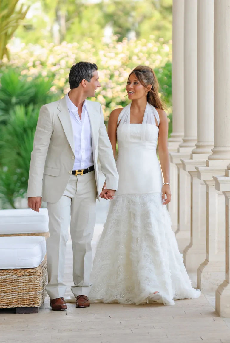 Bride and groom holding hands and walking together in a columned outdoor corridor surrounded by greenery.