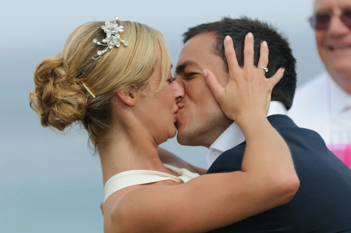 Bride and groom sharing a kiss during wedding ceremony, the bride's hand with a ring on the groom's face.