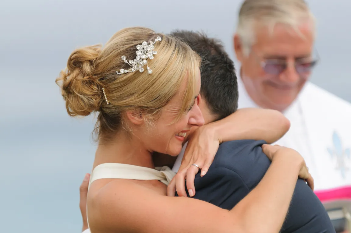 Bride with an embellished hairpiece embracing groom during outdoor wedding ceremony with officiant smiling in the background.