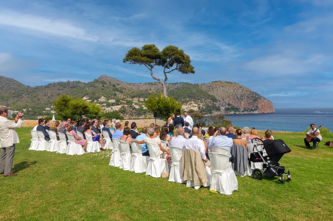 Outdoor wedding ceremony with guests seated on white chairs facing the ocean, mountains, and a guitarist playing nearby.
