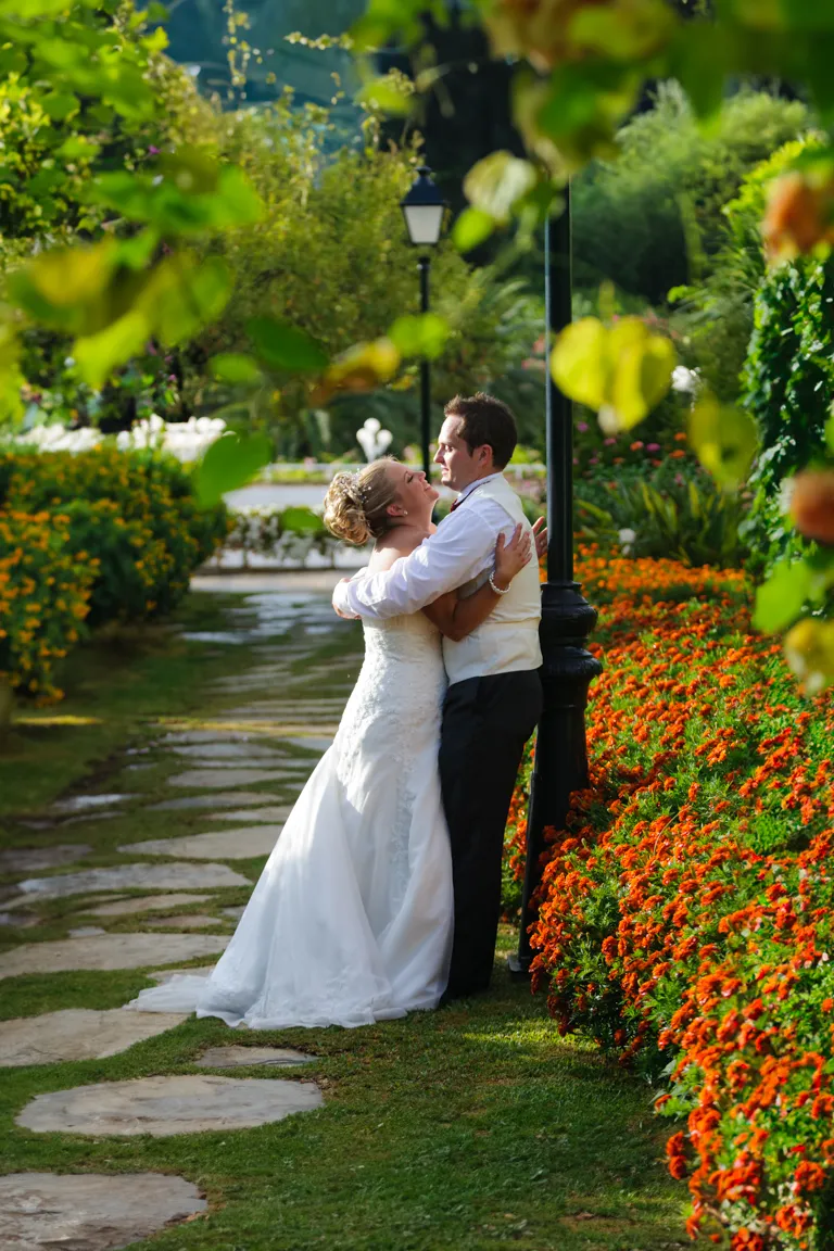 Bride and groom embracing in a garden with orange flowers along a stone pathway.