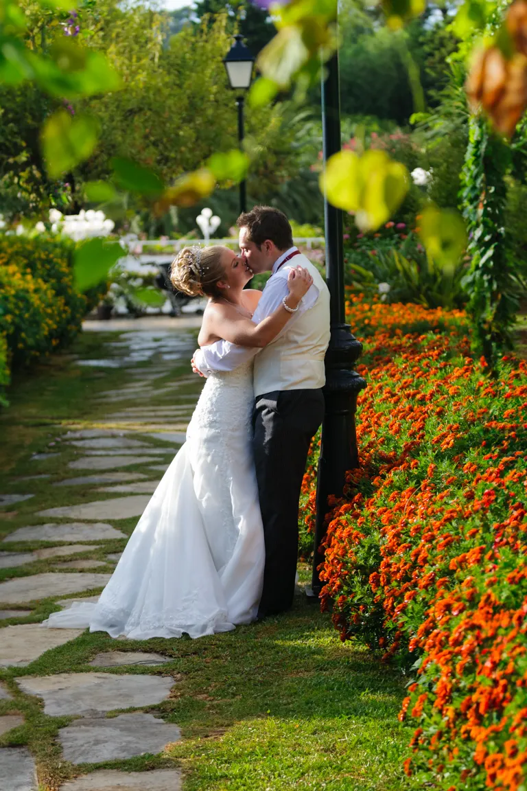 Bride and groom embracing and kissing on a garden path lined with vibrant orange flowers.