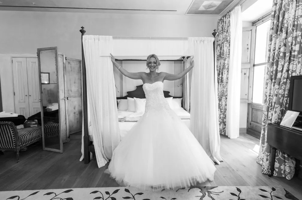 Smiling bride in strapless wedding dress holding open curtains of a four-poster bed in a bright, decorated room.