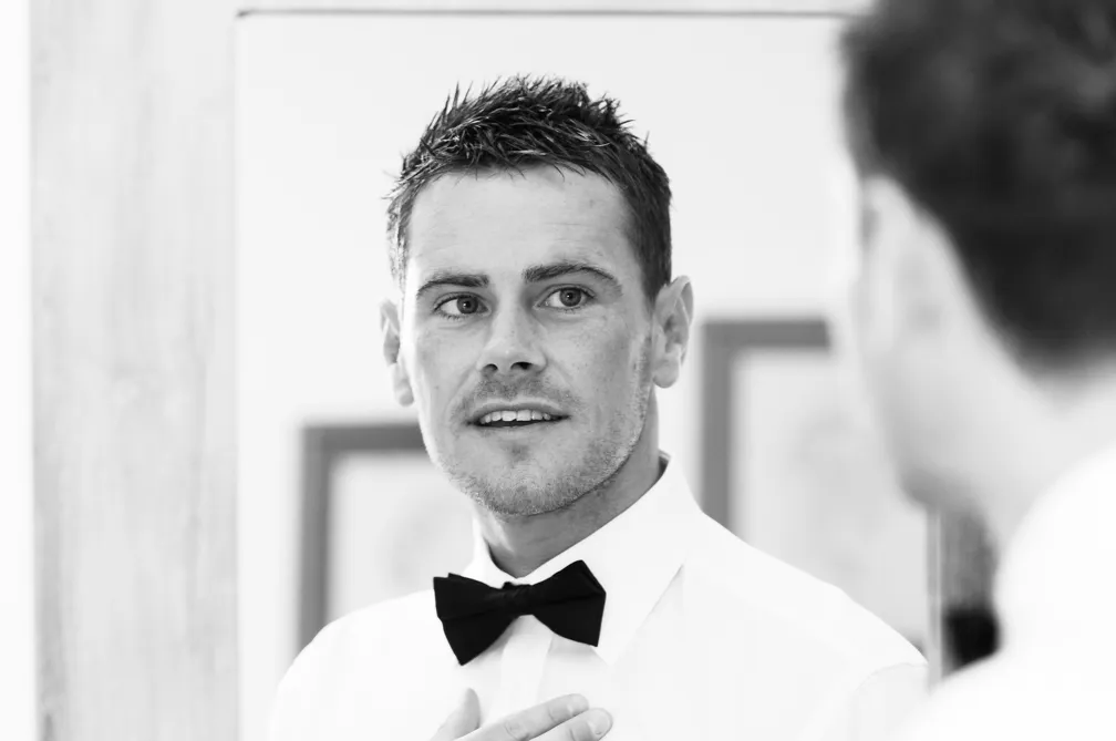 Man with short dark hair wearing a white shirt and black bow tie looking at himself in a mirror.
