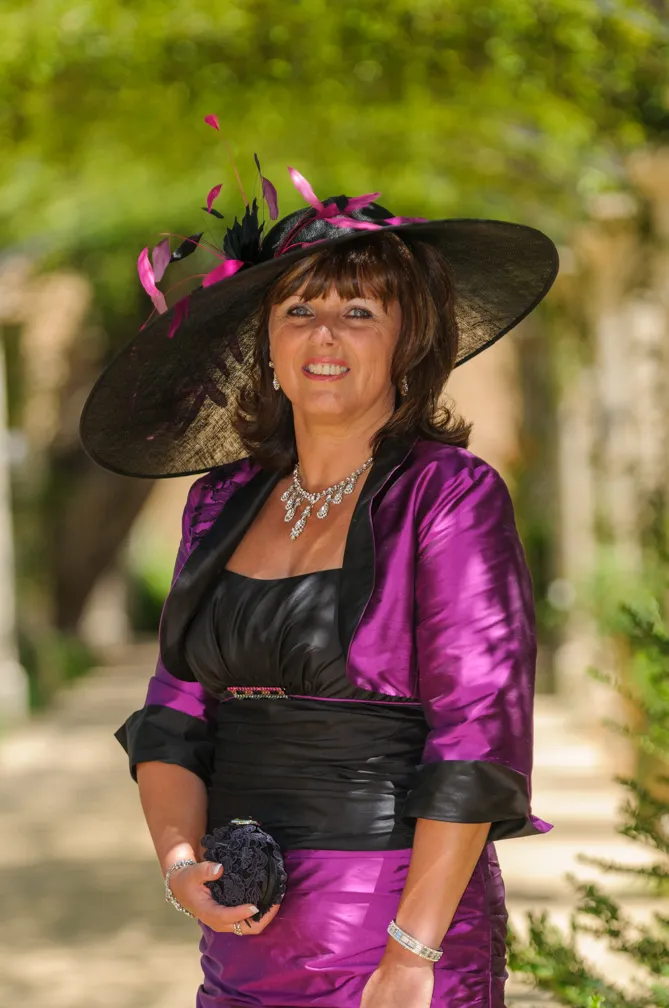 Woman wearing a purple satin dress and matching jacket with a large black hat adorned with purple feathers, holding a black clutch purse.