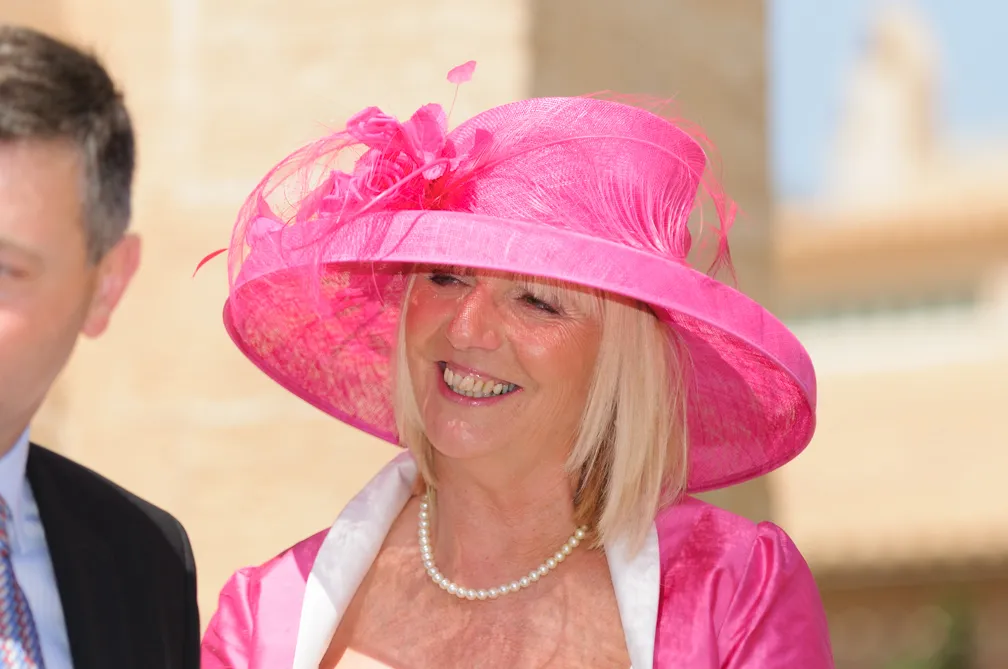 Smiling woman wearing a bright pink wide-brimmed hat with decorations and a matching pink outfit, with a man partially visible beside her.