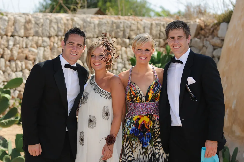Two men in black tuxedos and two women in colorful formal dresses posing outdoors in front of a stone wall.