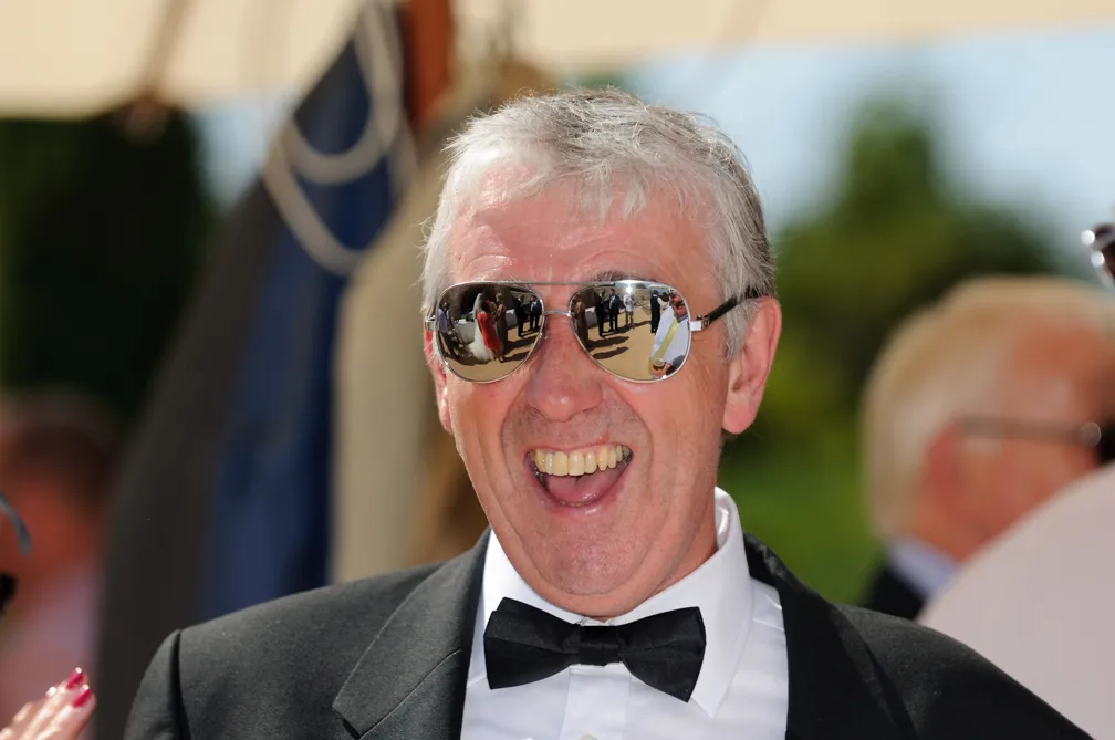 Smiling man in a tuxedo and black bow tie wearing reflective aviator sunglasses outdoors.