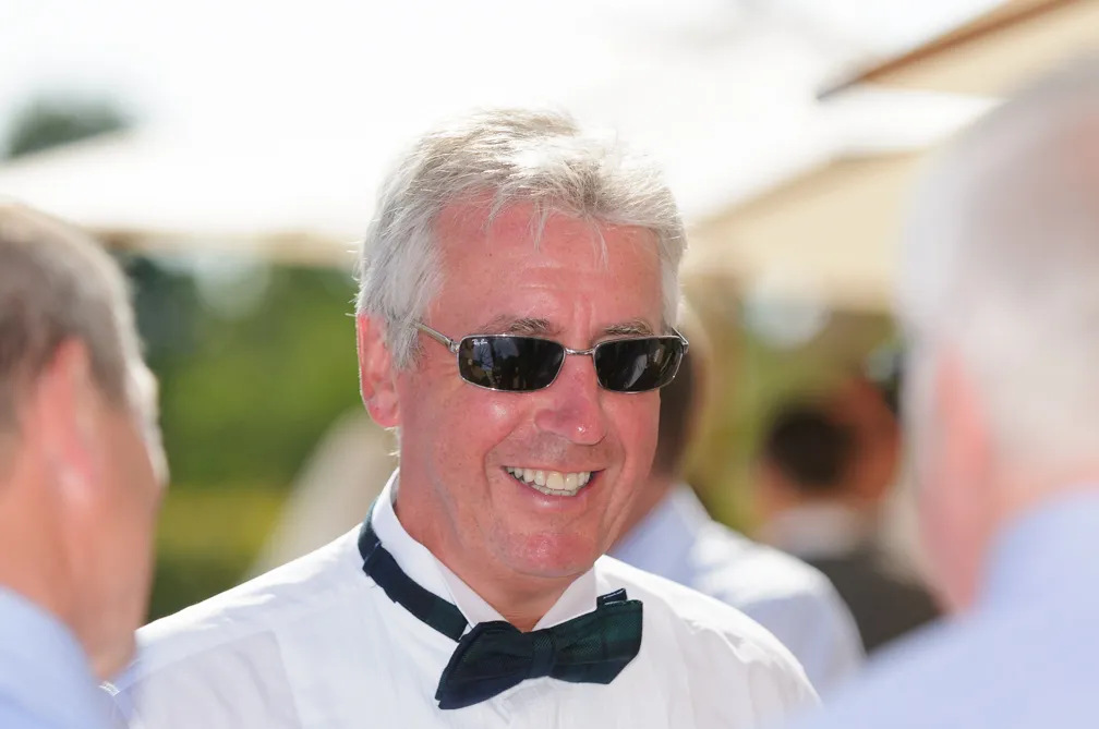 Smiling older man with gray hair wearing sunglasses, a white shirt, and a black bow tie at an outdoor event.