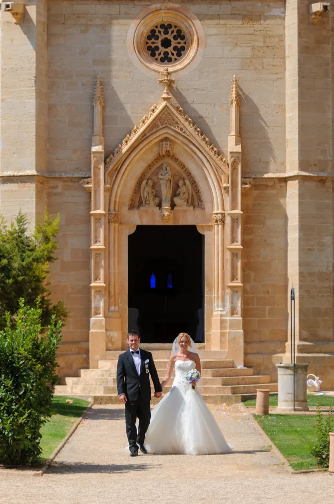 Bride and groom holding hands and walking on a path in front of a large stone church entrance with ornate carvings.