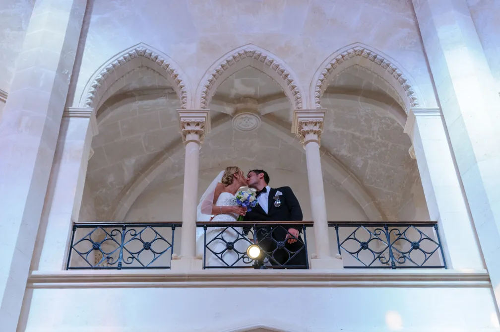 Bride and groom kissing on a balcony with stone arches in the background.