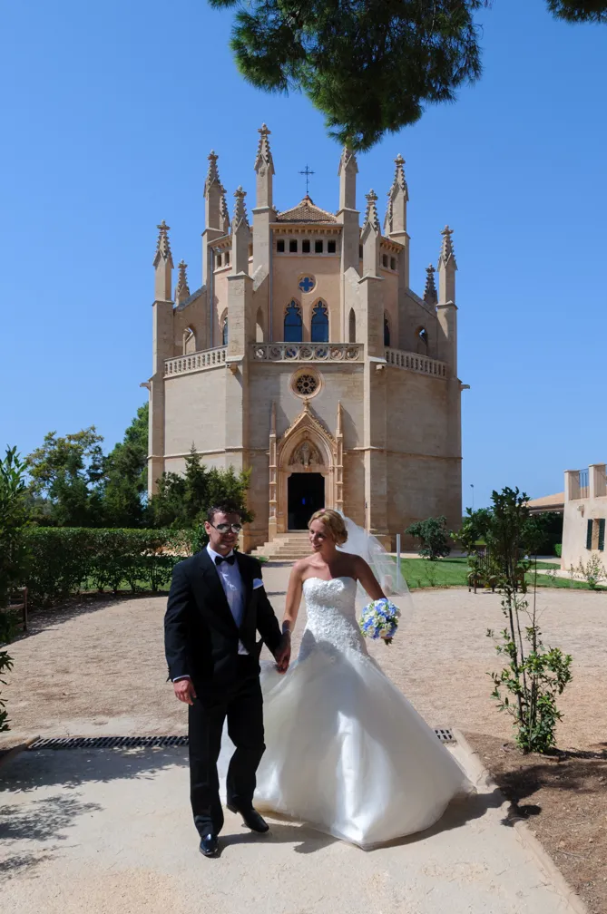Bride in a white wedding gown holding a bouquet and groom in a black tuxedo walking hand in hand outside a Gothic-style stone chapel.