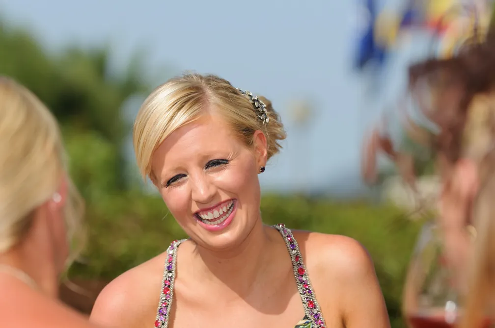 Blonde woman laughing outdoors in a decorative sleeveless dress.