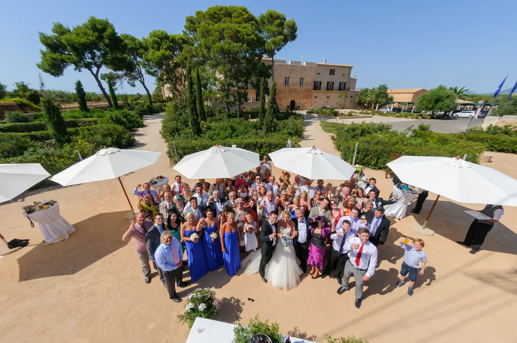 Large group of wedding guests gathered outdoors under white umbrellas with bride and groom in the center.