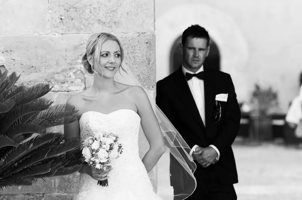 Bride holding a bouquet and smiling, standing near a stone wall with groom in a tuxedo blurred in the background.
