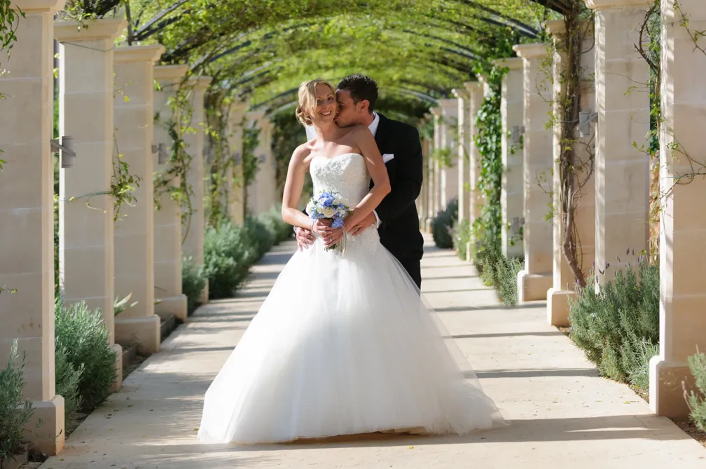 Bride in a white wedding dress holding a bouquet, embraced and kissed on the cheek by groom in a black suit under a leafy outdoor archway.