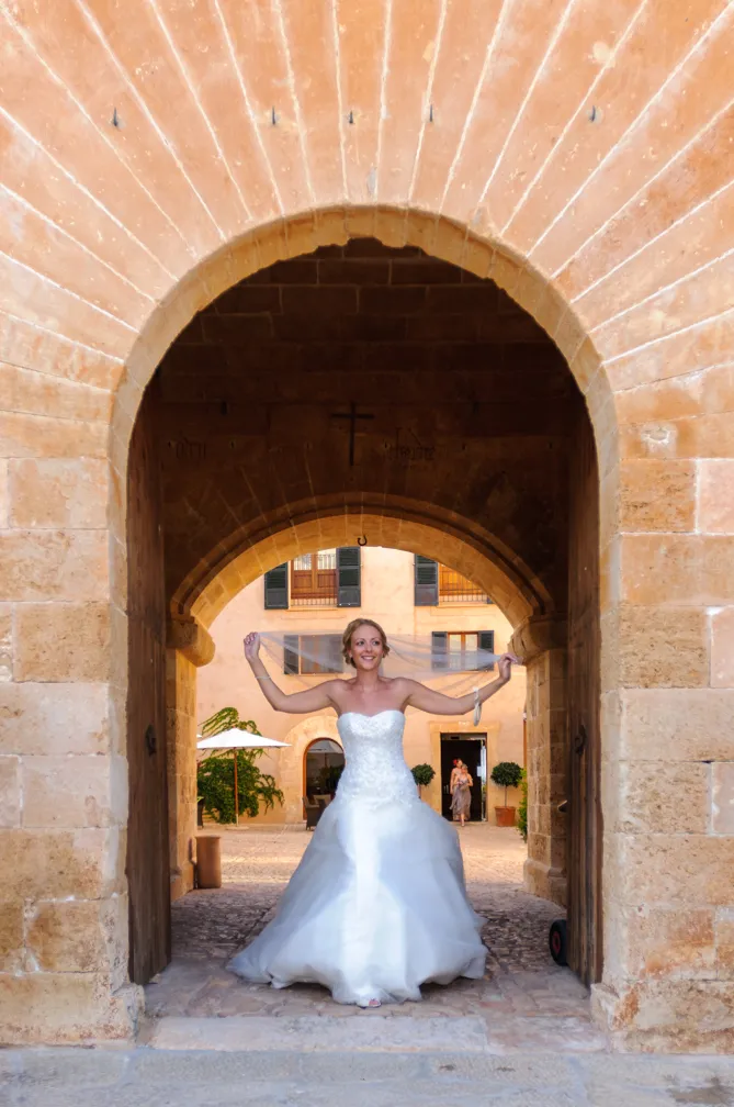Bride in a strapless white wedding gown standing under an arched stone doorway, holding her veil above her head.
