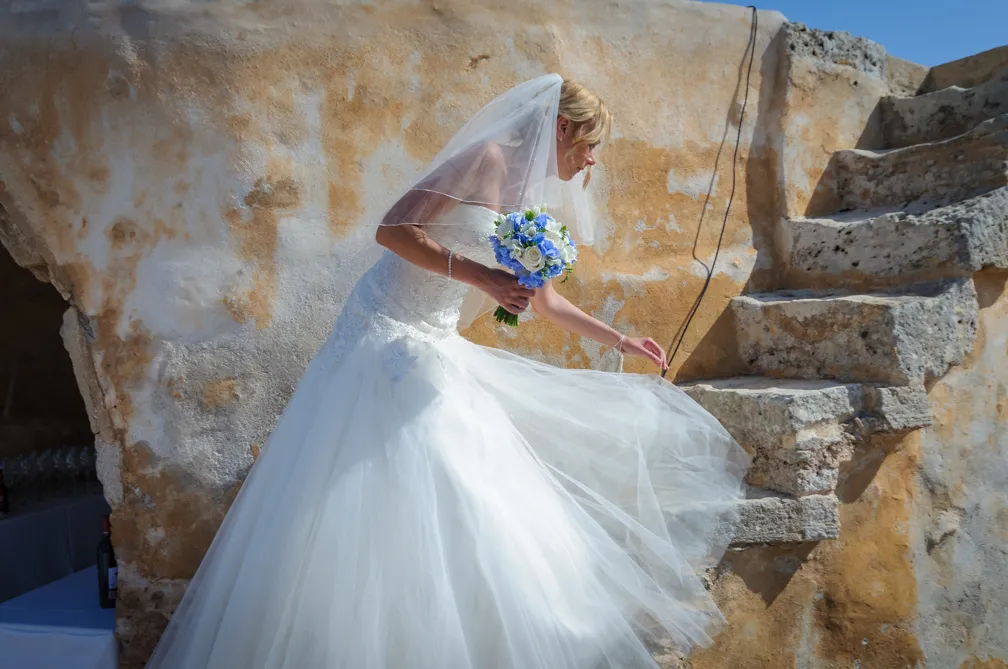 Bride in white wedding dress and veil holding blue and white bouquet, lifting her skirt while walking up rustic stone stairs.