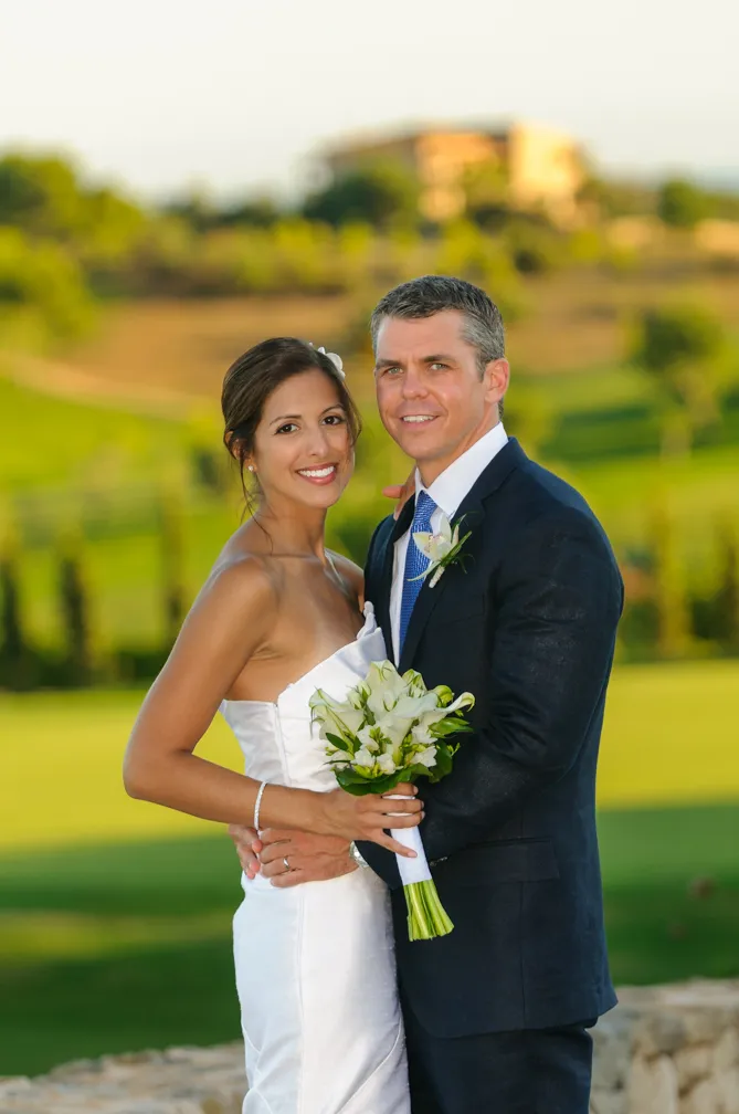 Bride in white dress holding a bouquet and groom in dark suit embracing outdoors with greenery in the background.