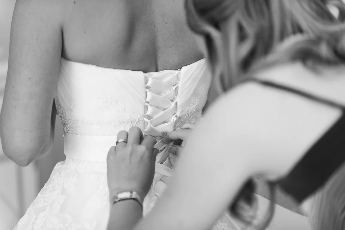 Close-up black and white photo of a woman lacing up the back of a bride's wedding dress.