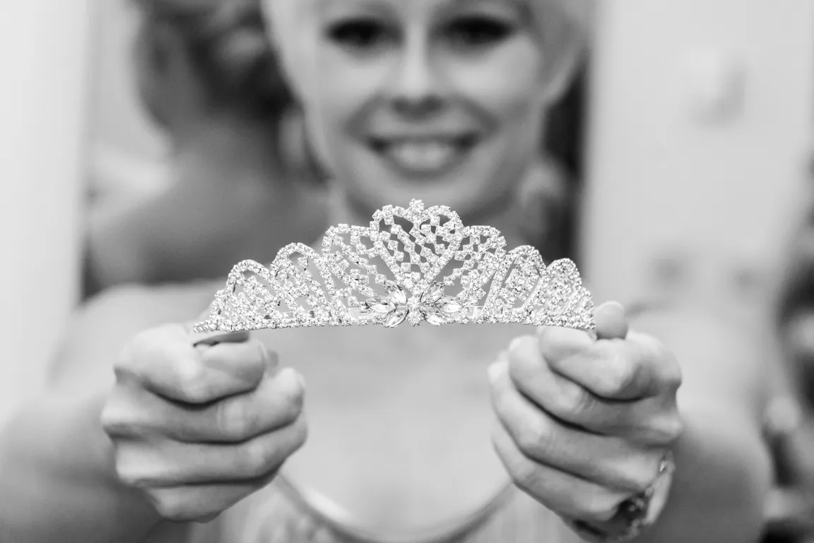 Woman smiling while holding a sparkling tiara towards the camera in a black and white photo.