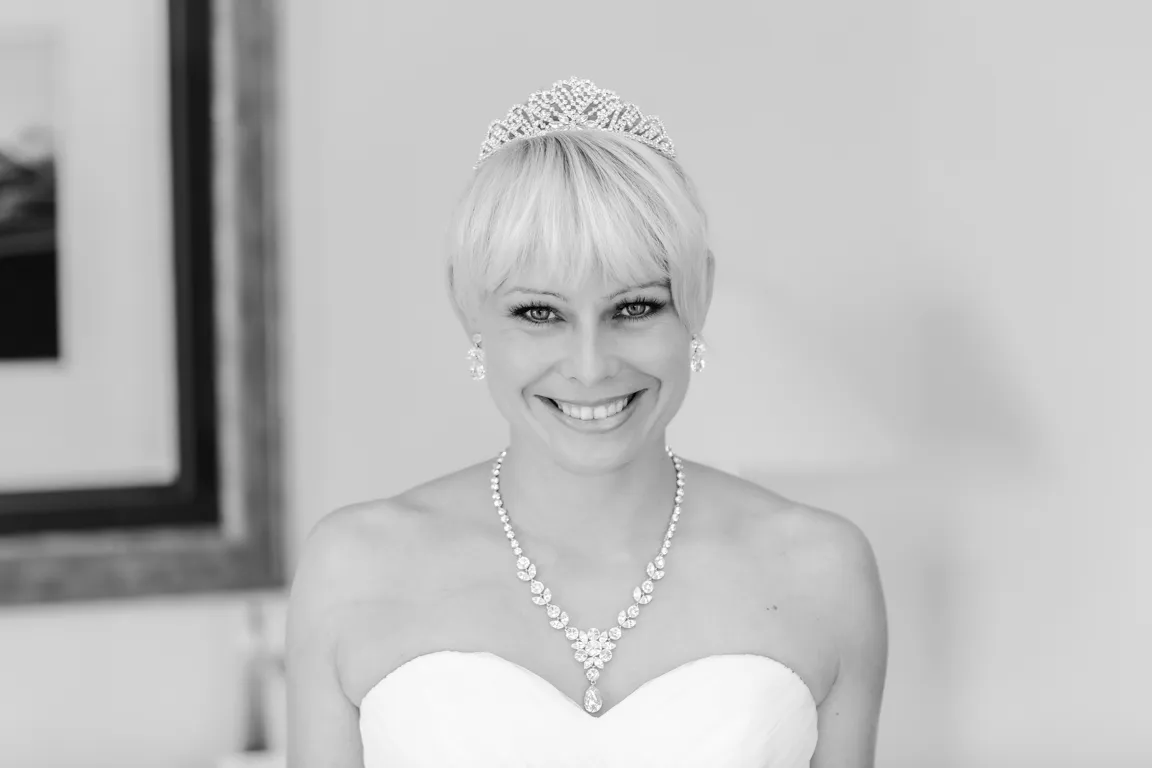 Smiling bride with short hair wearing a tiara, earrings, a necklace, and a strapless wedding dress.