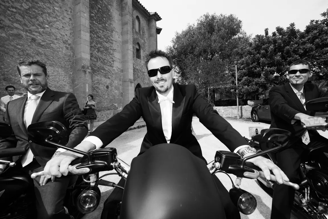 Three men in suits and sunglasses sitting on motorcycles outside near a stone building and trees.