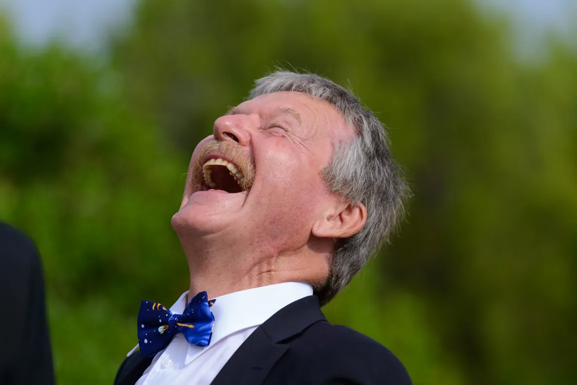 Middle-aged man with gray hair and mustache laughing heartily outdoors wearing a blue patterned bow tie and dark suit jacket.