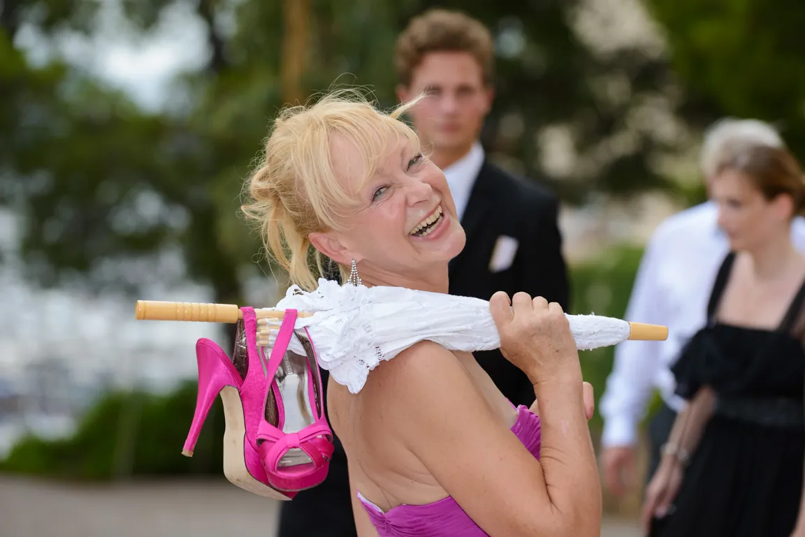 Smiling woman in a purple dress carrying white lace umbrella and pink high heels on her shoulder at an outdoor event.