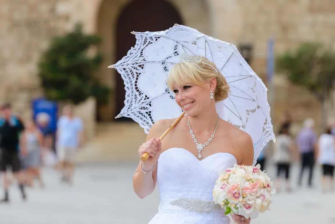 Smiling bride in white strapless wedding dress holding a lace parasol and bouquet of pink and white flowers.