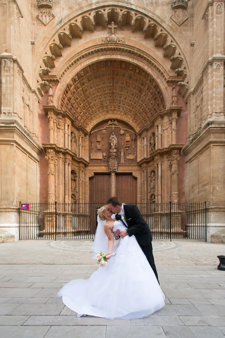 Bride and groom kissing in front of an ornate, large cathedral entrance with detailed stone carvings and wooden doors.