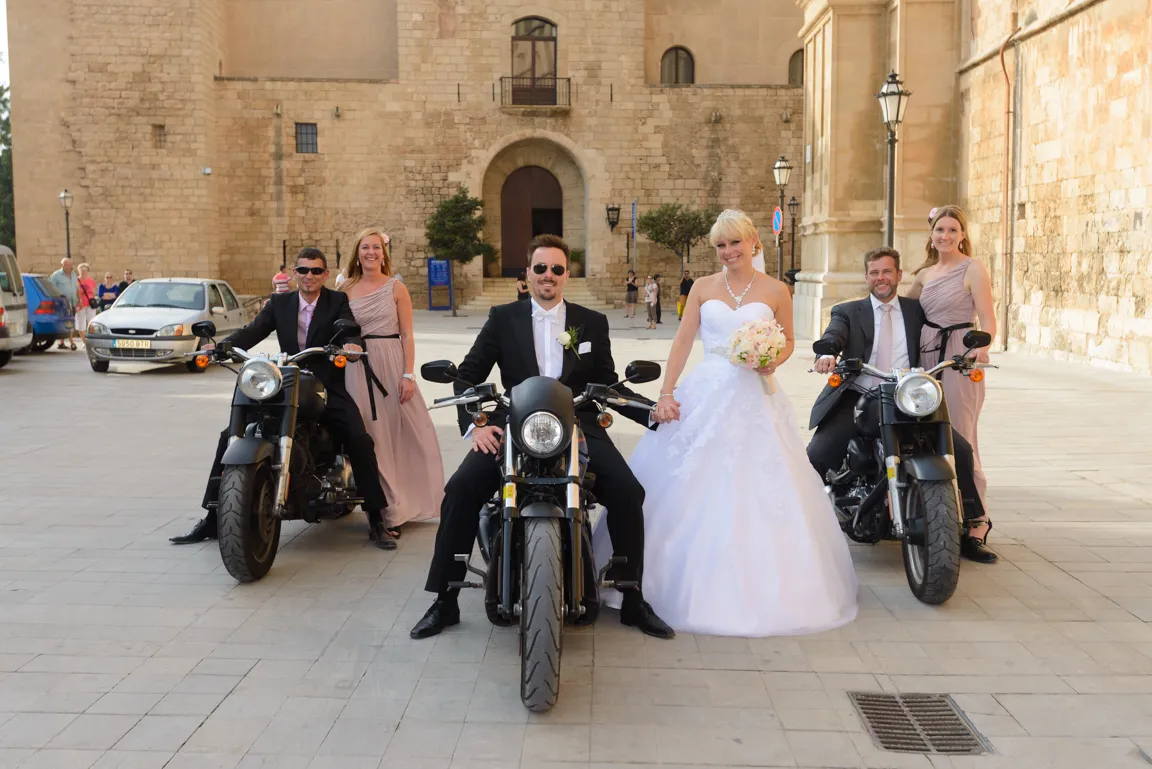 Bride in white wedding gown holding bouquet stands beside groom in tuxedo seated on motorcycle, flanked by two bridesmaids and groomsmen on motorcycles in a sunlit courtyard.