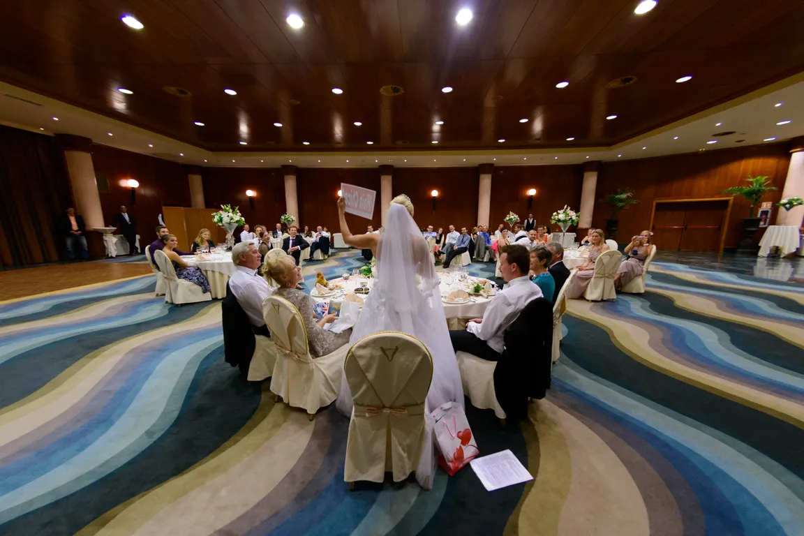 Bride in white wedding dress holding a sign while standing at a banquet table surrounded by seated guests in a large decorated hall.