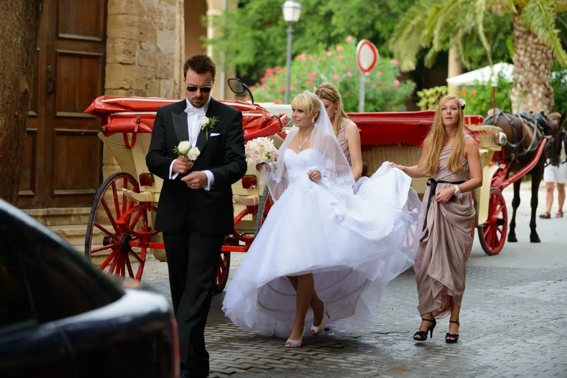 Bride in white wedding gown walking on a cobblestone street with bridesmaids and a groom in a black tuxedo holding white roses, passing by a red horse-drawn carriage.