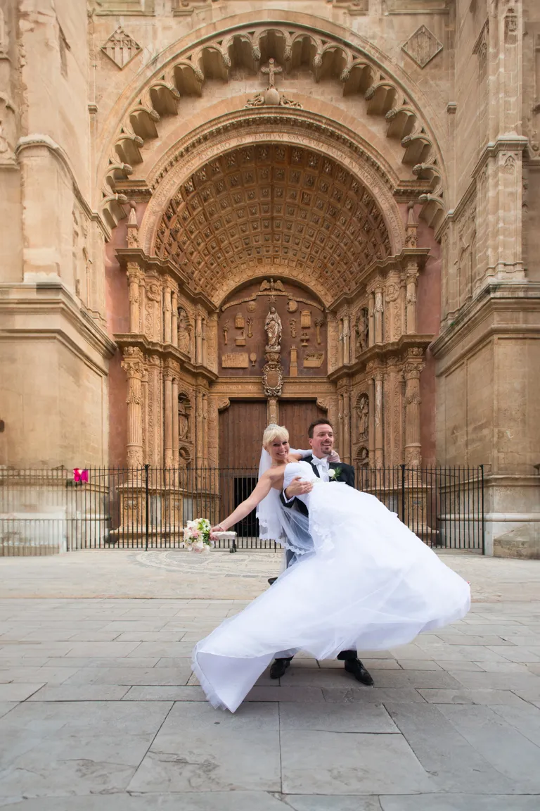 Groom lifting bride in a white dress and veil in front of an ornate historic church entrance.
