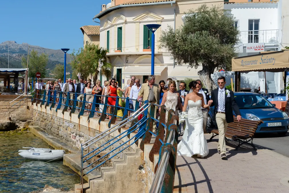 Bride and groom walking hand in hand along a waterfront promenade followed by a large group of wedding guests on a sunny day.