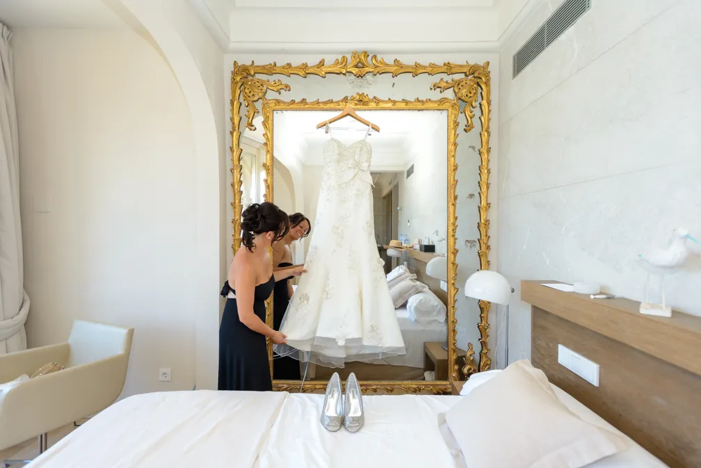 Two women in black dresses adjusting a white wedding gown hanging on a large ornate gold mirror in a bedroom.