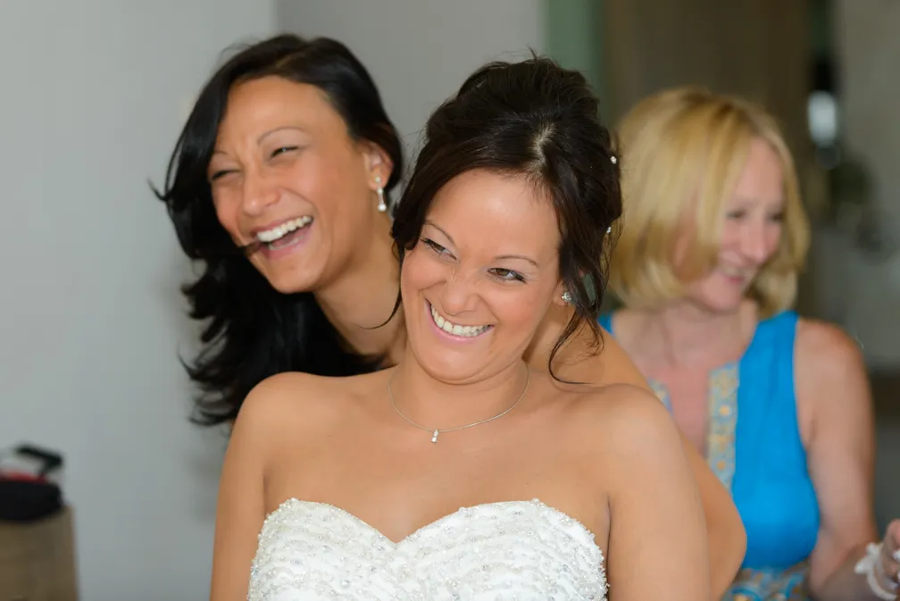 Bride in a white beaded strapless dress smiling with two women laughing behind her.