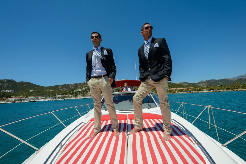 Two men in blazers, dress shirts, and khaki pants standing barefoot on the bow of a boat with a red and white striped cushion, overlooking turquoise water with mountains in the background.