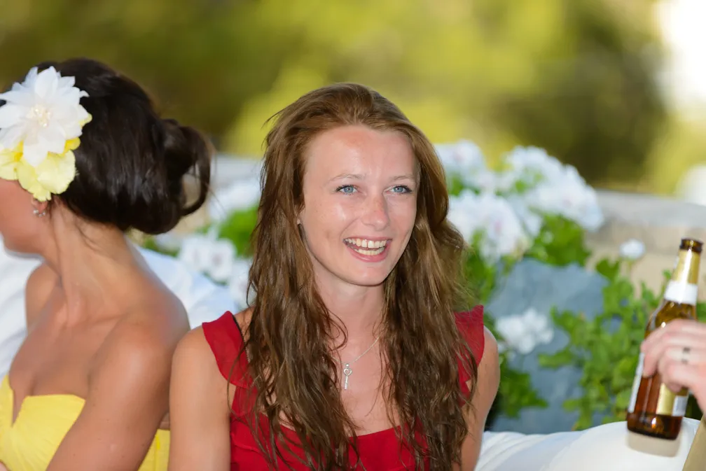 Smiling woman with long brown hair in a red dress sitting outdoors, another woman in a yellow dress with a white and yellow flower in her hair beside her.