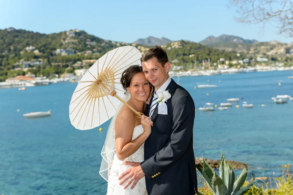 Bride in white dress holding a white parasol embraces groom in dark suit by a waterfront with boats and hills in the background.