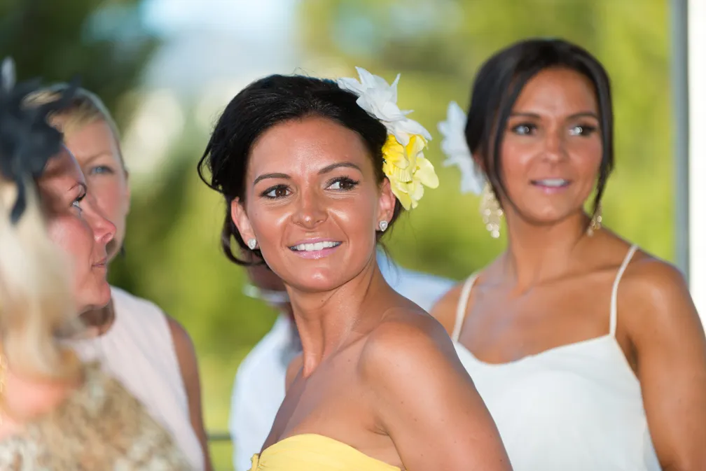 Close-up of a smiling woman in a yellow strapless dress with white and yellow flowers in her dark hair, surrounded by other women in light-colored dresses.