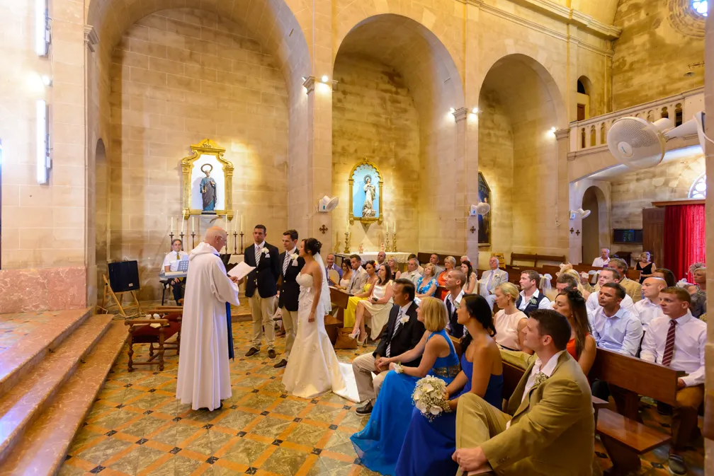 Bride and groom standing before a priest in a church ceremony with guests seated in pews watching.
