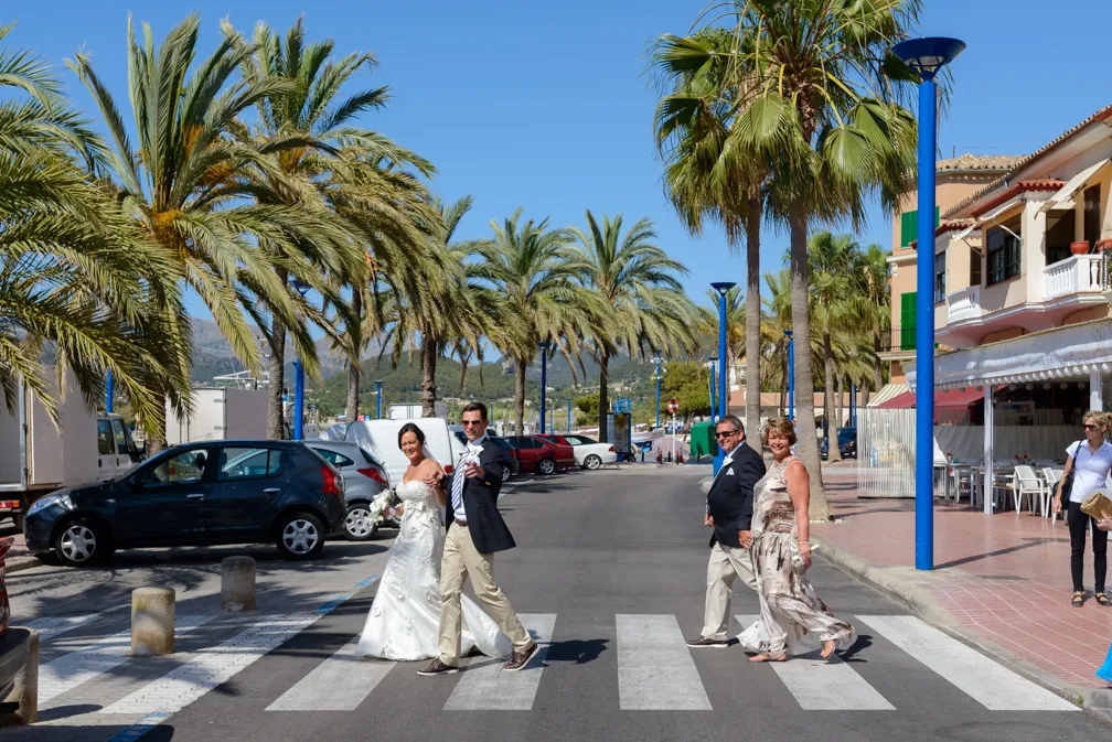 A bride and groom in wedding attire and two formally dressed guests crossing a palm-lined street on a sunny day.